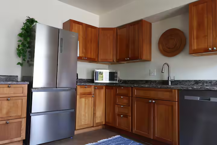 Kitchen with wooden cabinets, a stainless steel refrigerator, a microwave on the countertop, a sink with a faucet, and a decorative wall hanging.