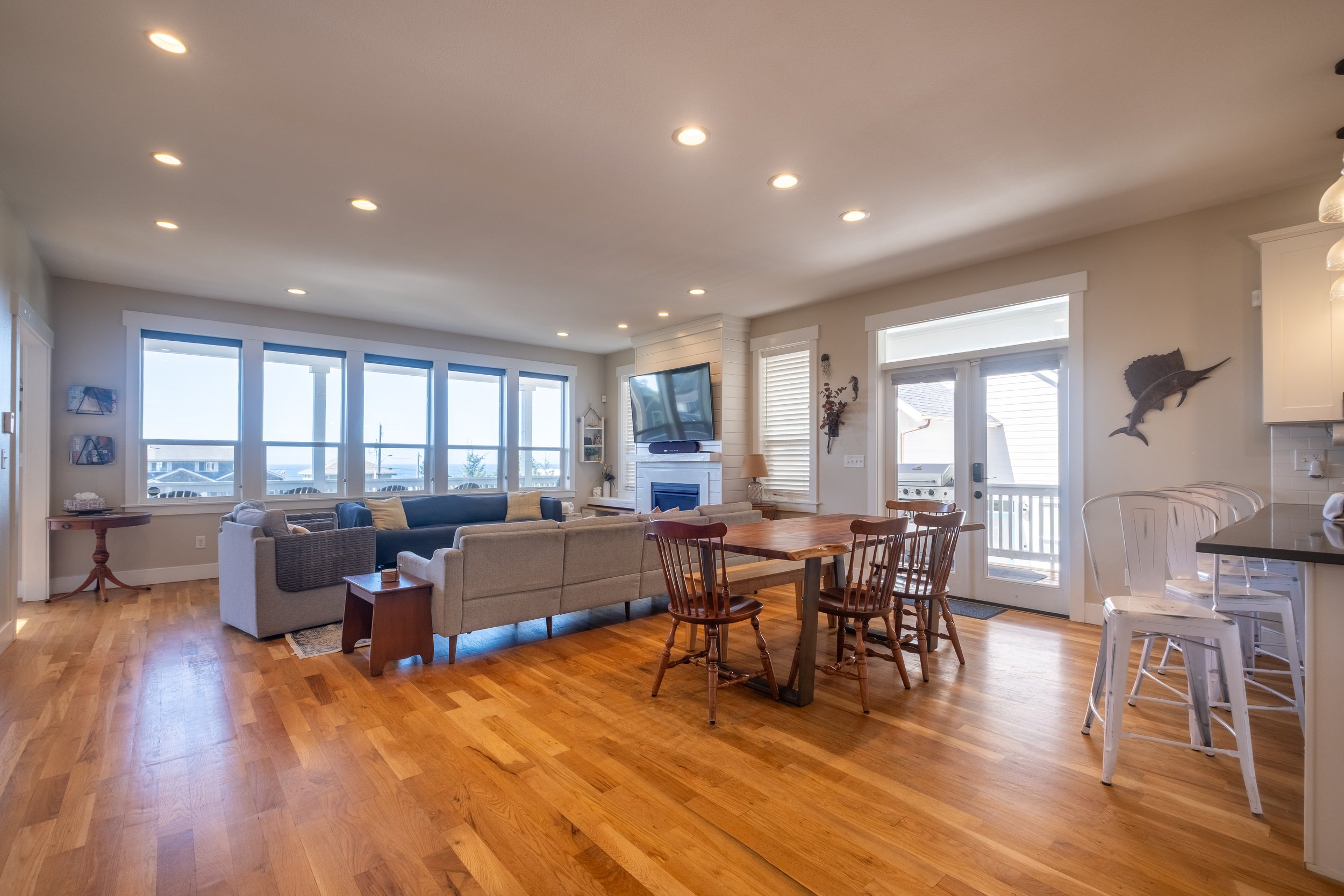 Living room with hardwood floors, beige couches, a wooden dining table with chairs, large windows with blinds showing outdoor view, a television on a white fireplace mantel, and decorative wall art including a fish. Kitchen area partially visible wit