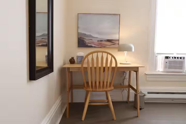 A wooden desk with a matching wooden chair in front of a beige wall decorated with an abstract landscape painting. A table lamp is on the desk, and there is an air conditioning unit in the window to the right.