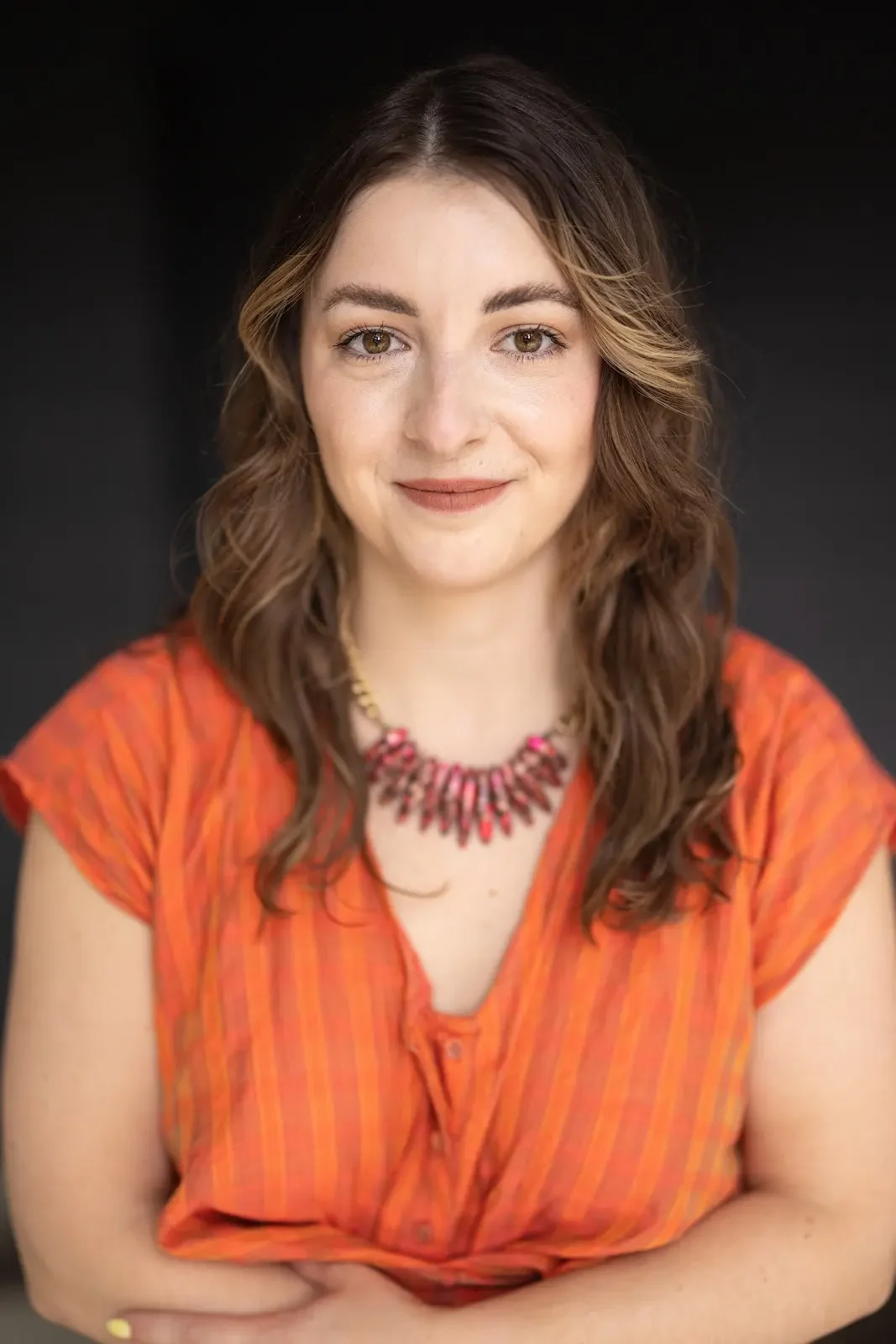 Young woman with shoulder-length wavy brown hair, wearing an orange top with a necklace, smiling against a black background.