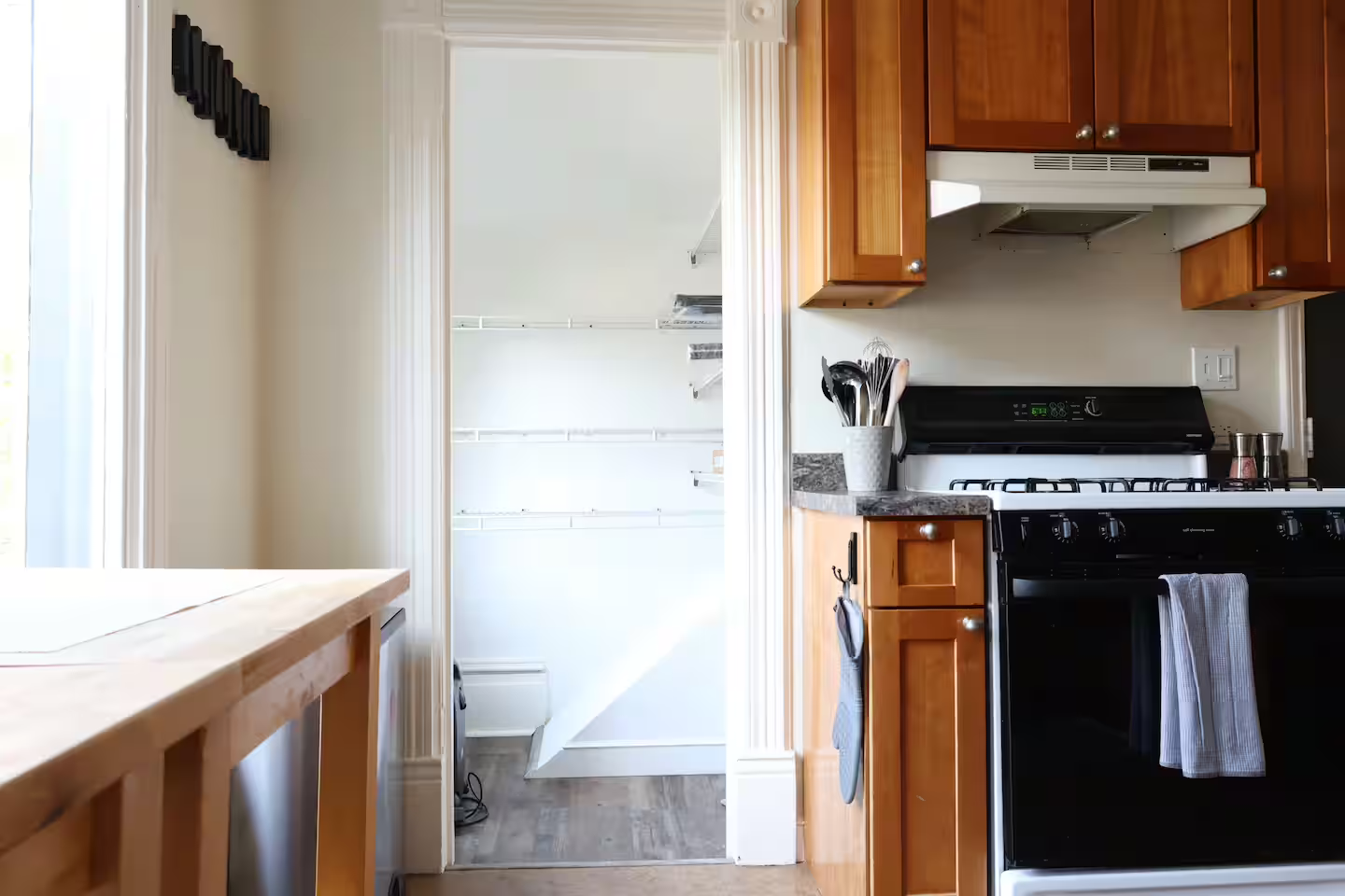 Kitchen with wooden cabinets, black gas stove with a towel hanging, a countertop with utensils, and a walk-in pantry with shelves in the background.