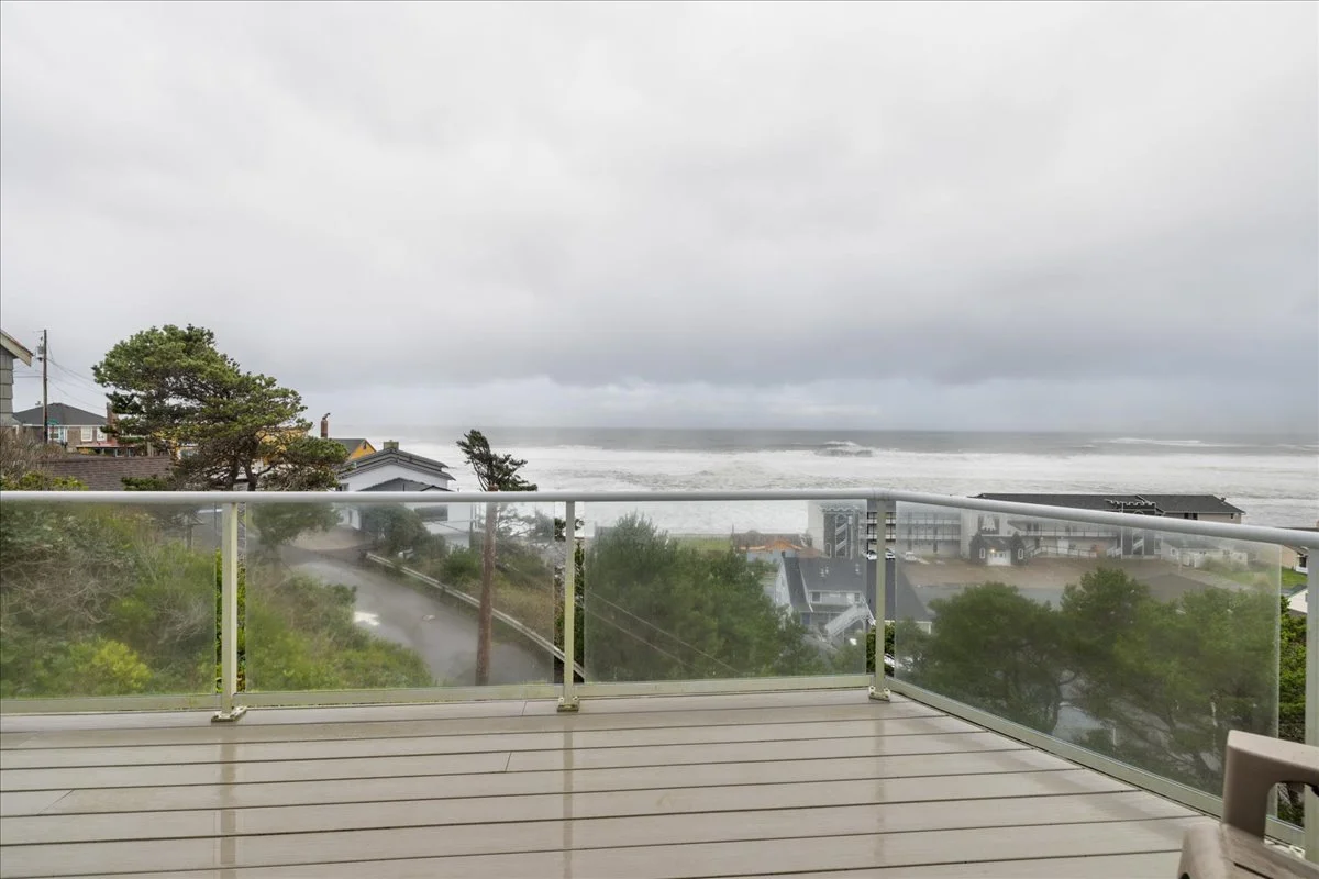 View from a balcony showing a cloudy sky, ocean waves, trees, and houses on a coastline.