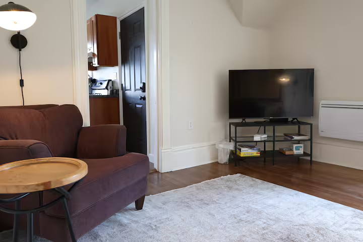 Living room with brown sofa, side table, wall-mounted lamp, flat-screen TV on black stand, white area rug, hardwood floors, and a glimpse of a kitchen through an open door.