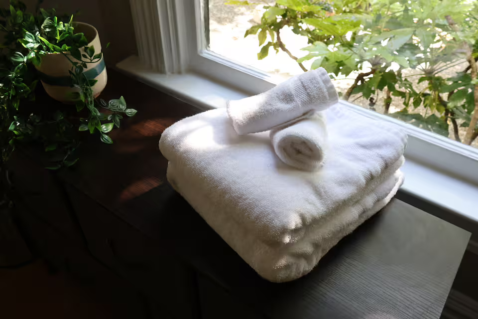 Stacked white towels on a dark wooden surface by a window, with a potted plant on the left and leafy greenery visible outside.