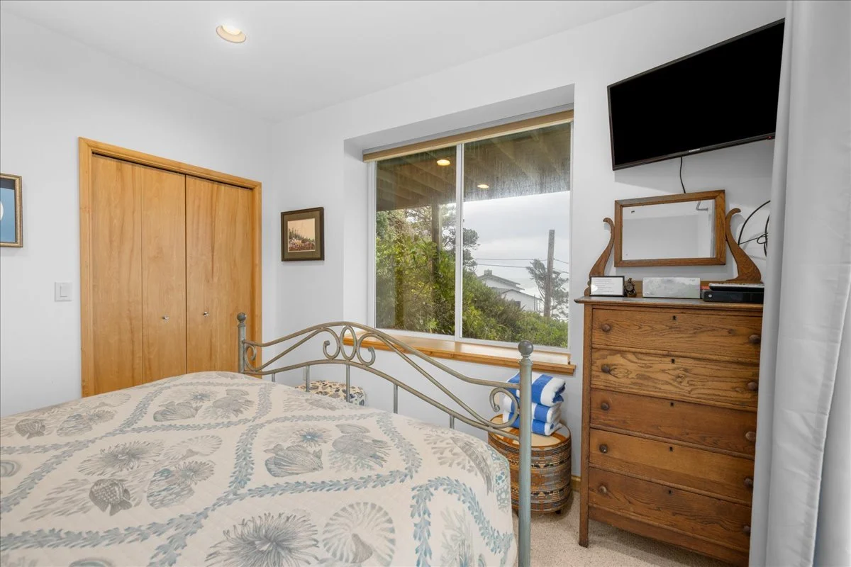 A bedroom with a metal bed frame, a wooden dresser with a mirror, a television mounted on the wall, a window with a view of trees and houses, and a closet with wooden double doors.