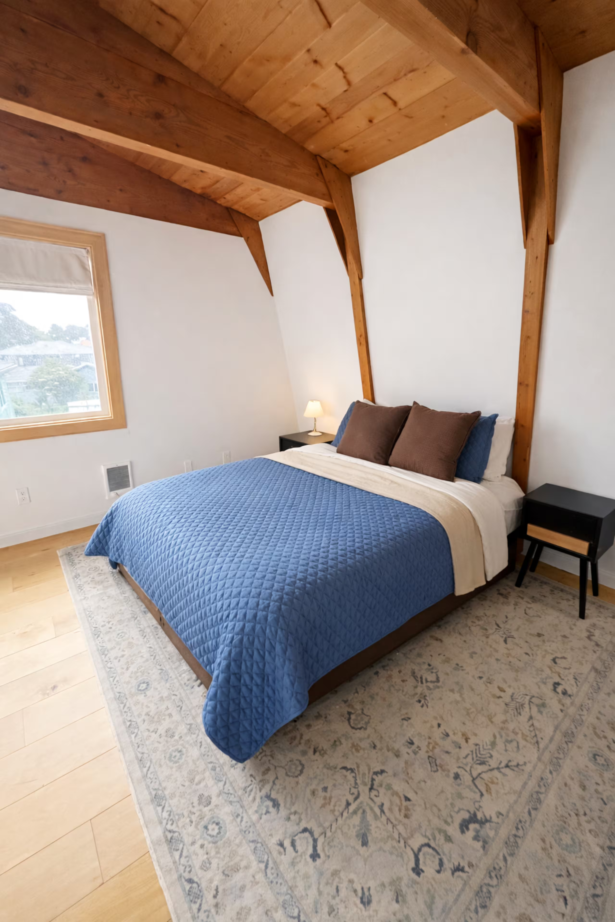 A bedroom with a bed covered in a blue quilt, two brown pillows, and two navy blue pillows. There are black nightstands on each side of the bed, with a lamp on the right. The room has a window with a beige blind, white walls, wooden ceiling beams, an