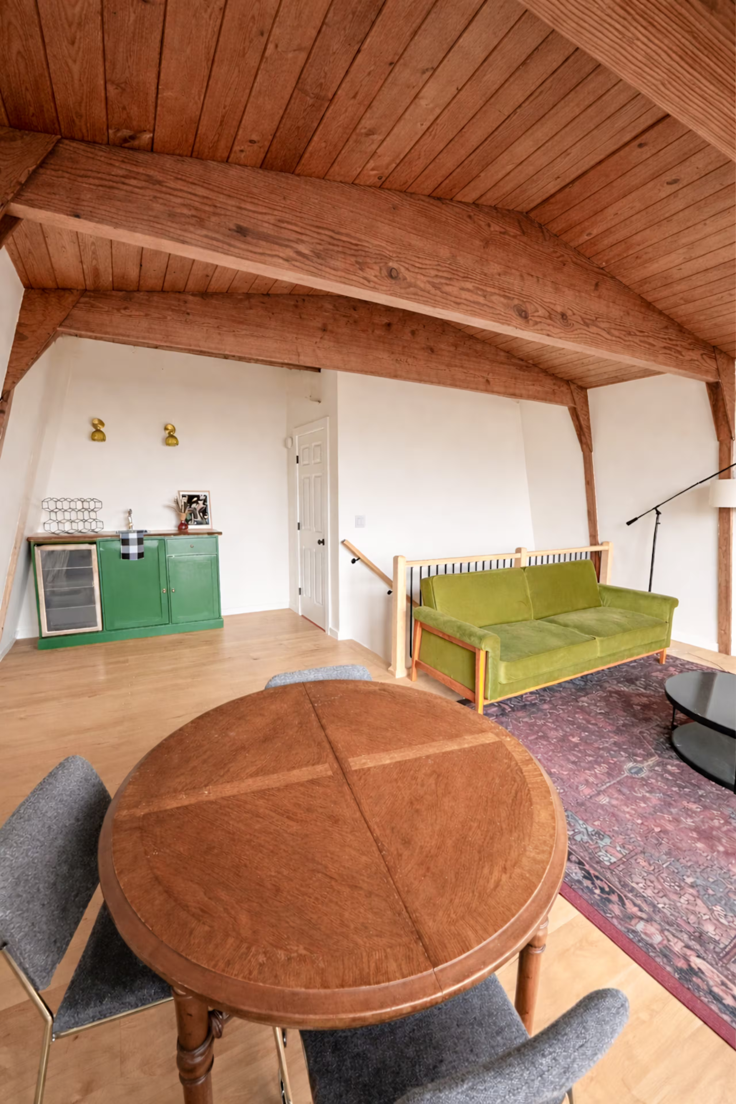 A round wooden dining table with four chairs, two upholstered with dark grey fabric and two with patterned fabric, in a living room with a wooden ceiling, a green sofa, a standing lamp, and a Persian rug.