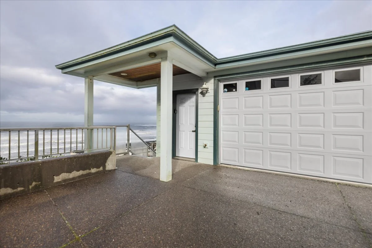 A house with a garage door and a front door located on a concrete patio with a view of the ocean in the background, under cloudy sky.