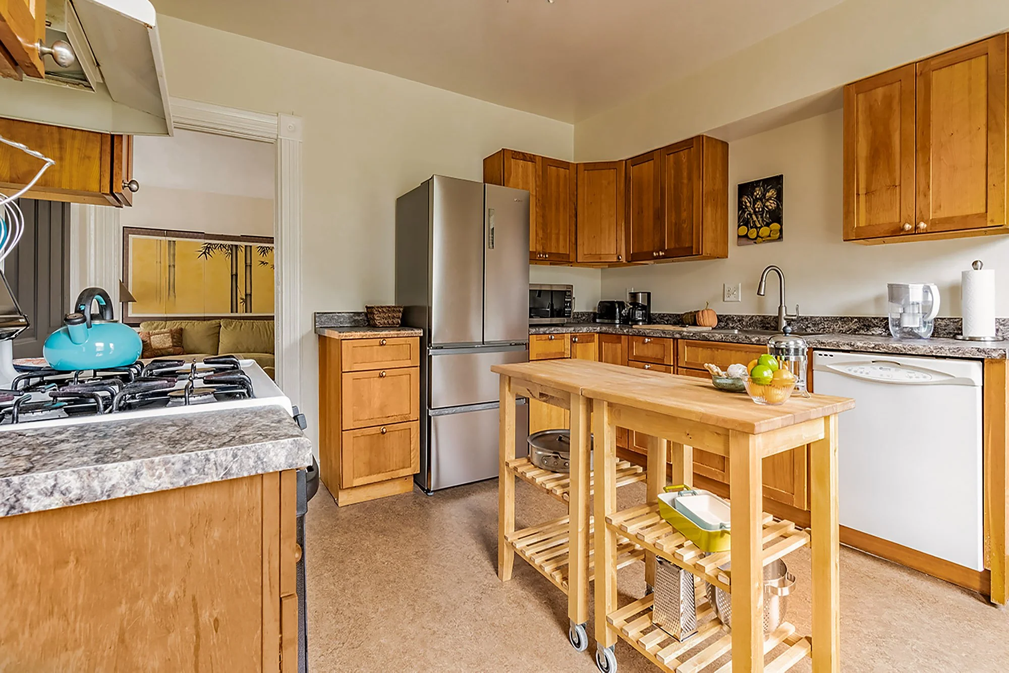 A kitchen with wooden cabinets, a stainless steel refrigerator, a microwave, coffee maker, and a pumpkin on the countertop. A wooden island in the center has a storage shelf and a cutting board on top. A bowl of apples and a bottle of water are on the island, with a paper towel roll on the counter. Part of a stove with a blue kettle is visible in the foreground. There is a doorway leading to a living room with a beige couch and a decorative room divider with a bamboo design.