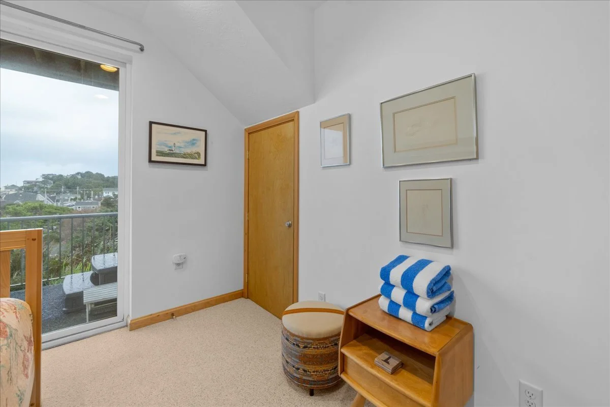 A corner of a bedroom with a sliding glass door leading to a balcony, a wooden door, framed pictures on the wall, a small wooden shelf with folded blue and white towels, and a striped upholstered ottoman.