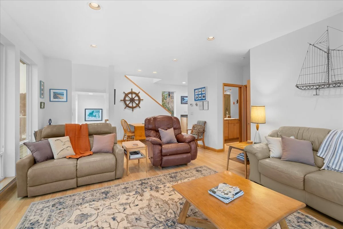 Living room with two beige sofas, one brown leather recliner, wooden coffee tables, a floor lamp, and nautical decor, including a ship's wheel and a wall-mounted ship model, with a large window and framed pictures on the walls.