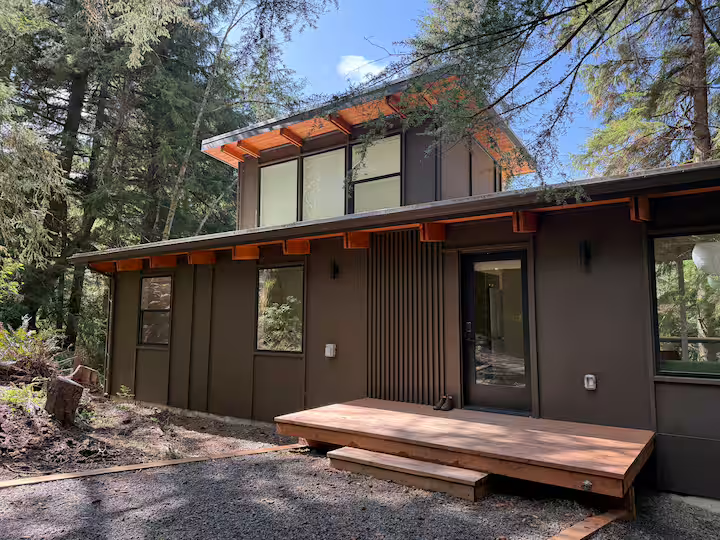 Modern two-story house surrounded by tall trees, with dark exterior walls, large windows, and a small wooden deck in front.