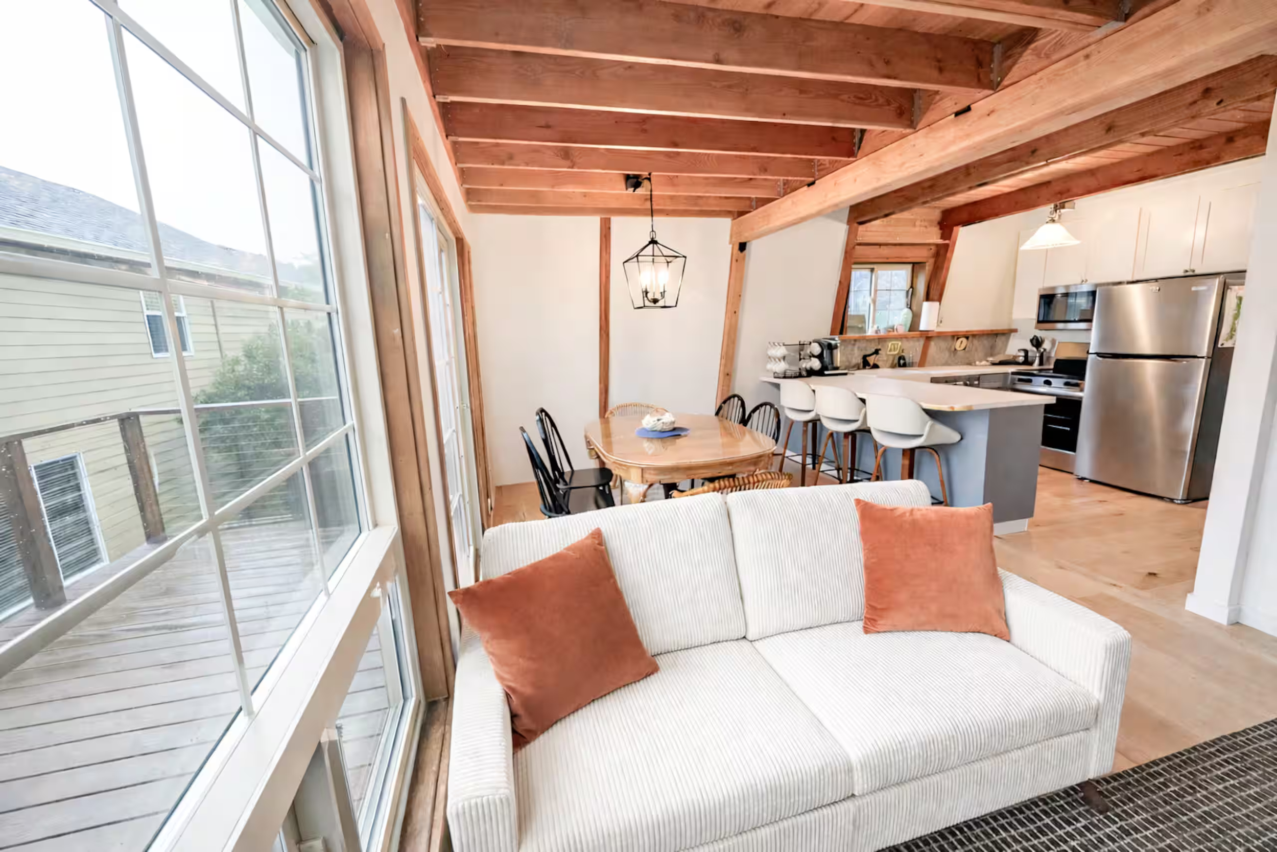 Living room with large windows, wooden ceiling beams, dining table with chairs, white couch with orange pillows, and a kitchen area with stainless steel appliances.