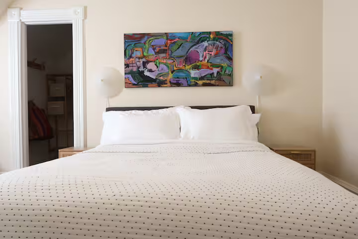 A neatly made bed with white bedding and pillows in a bedroom with a colorful abstract painting above it, beige walls, and a doorway to a closet.