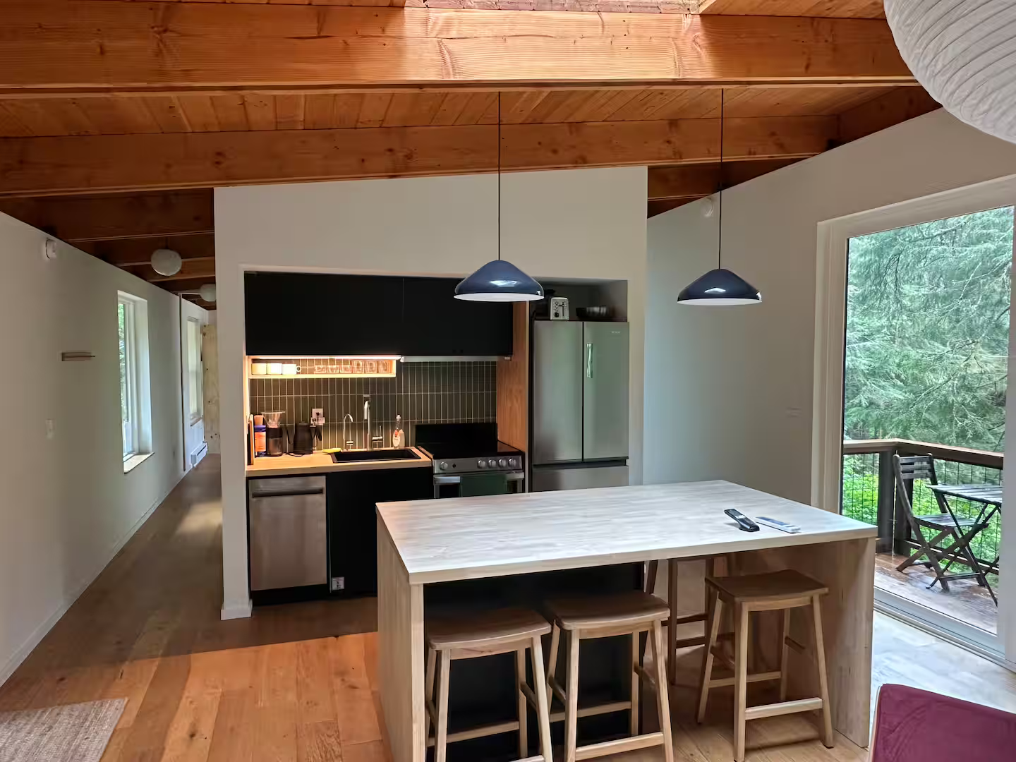 Kitchen with wooden ceiling, black and white cabinets, small black stove, stainless steel refrigerator, wooden dining table with stools, and sliding glass door leading to a balcony with outdoor chairs and greenery.