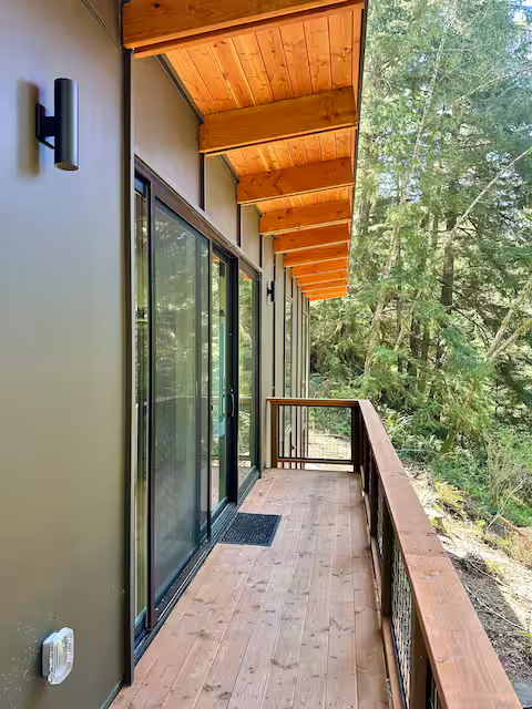 A balcony with wooden flooring and railing, attached to a house with sliding glass doors, surrounded by tall trees.