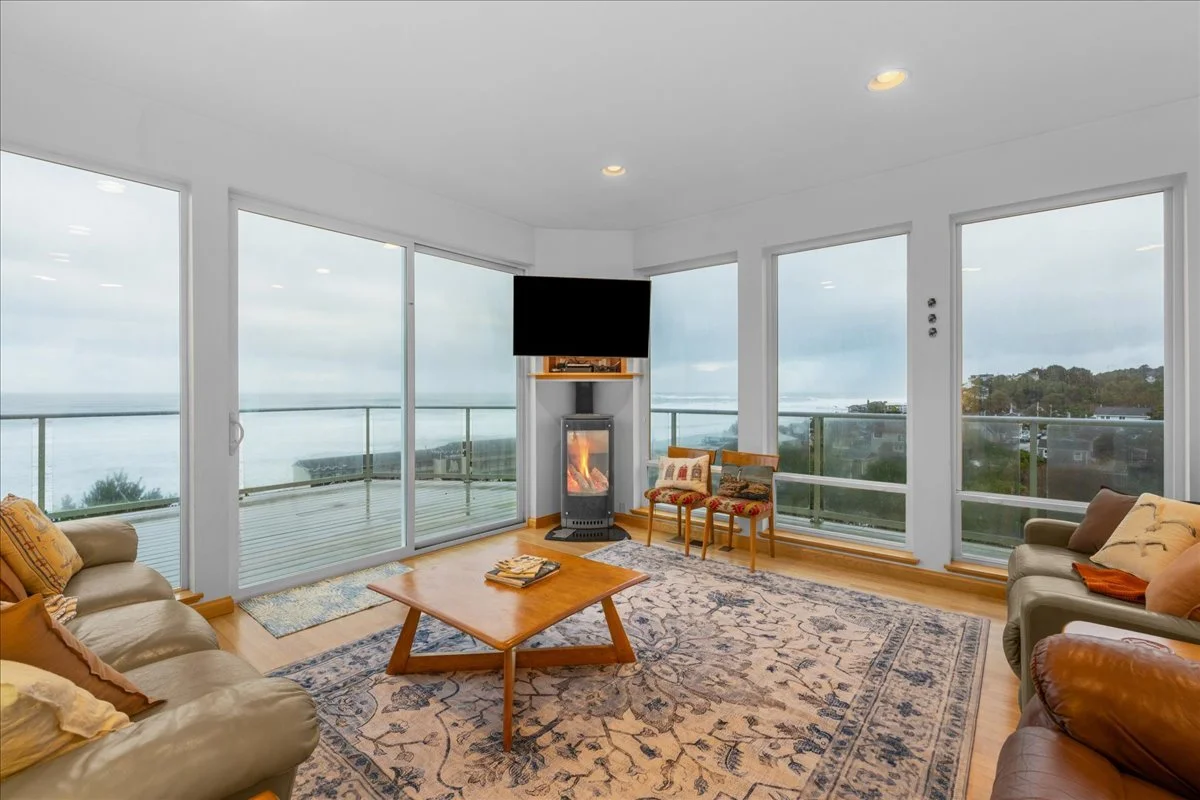 Living room with large windows overlooking a body of water and a balcony, a TV mounted above a fireplace, beige and brown couches, a wooden coffee table with magazines, a patterned rug, and two chairs near the windows.