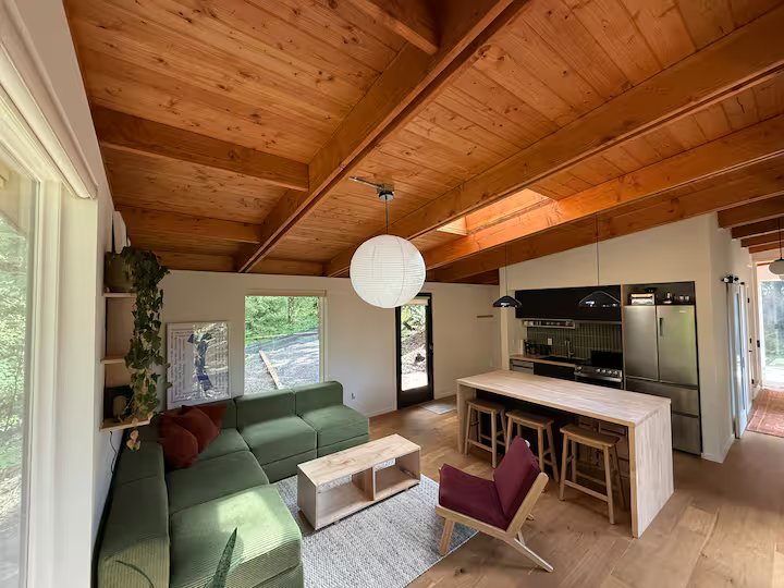 Interior of a living room and kitchen area with wooden ceiling, green sectional sofa, small wooden coffee table, armchair, kitchen island, and black cabinets.