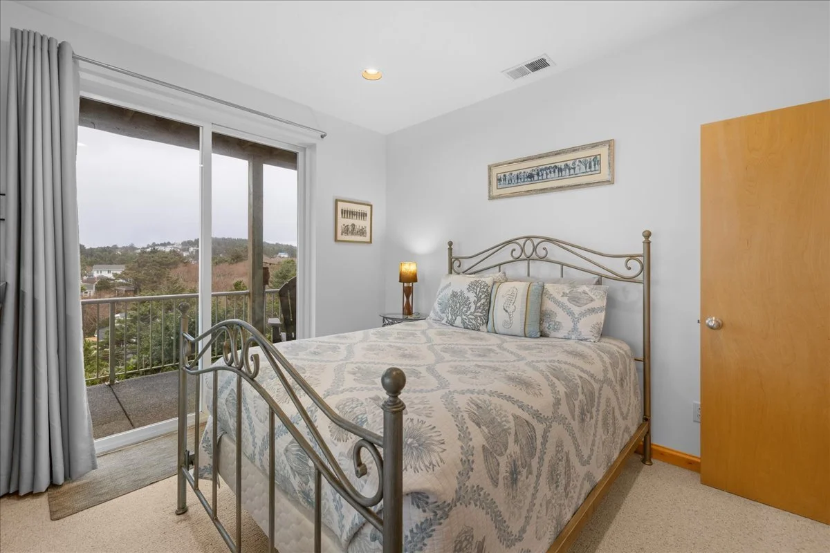 Bedroom with a bed, nightstand, lamp, framed artwork, and sliding glass door leading to a balcony with a view of trees and houses.