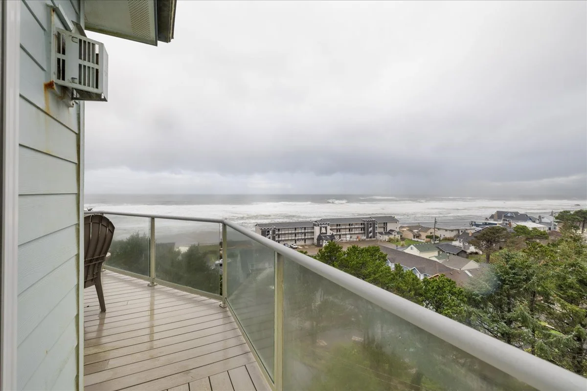 View from a balcony overlooking a coastal town with houses and trees, and the ocean with waves in the background under a cloudy sky.
