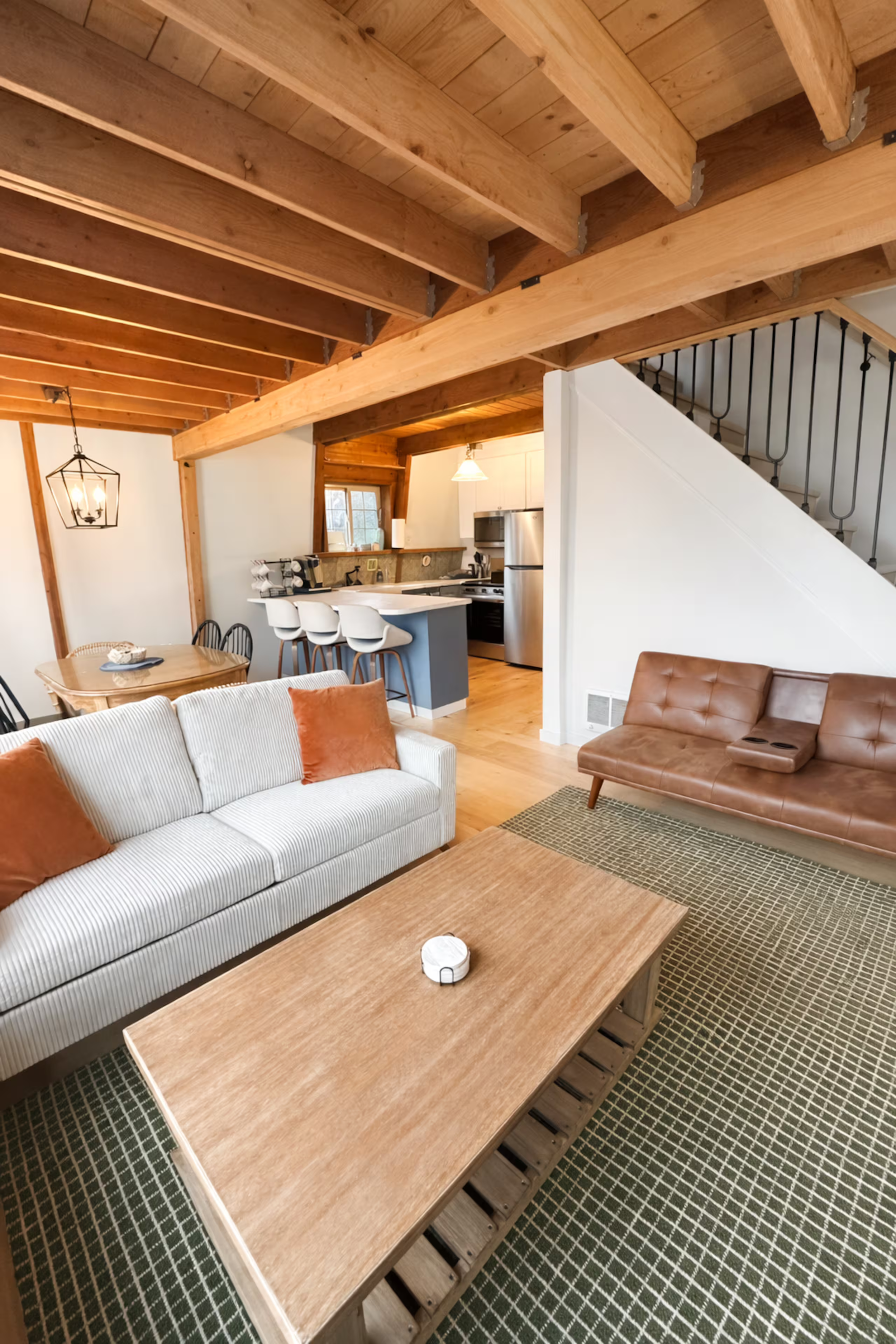Living room with wooden ceiling beams, a white couch with orange pillows, a wooden coffee table with a small device on it, a brown leather couch, a patterned rug, open kitchen with white cabinets, stainless steel appliances, a breakfast bar with whit