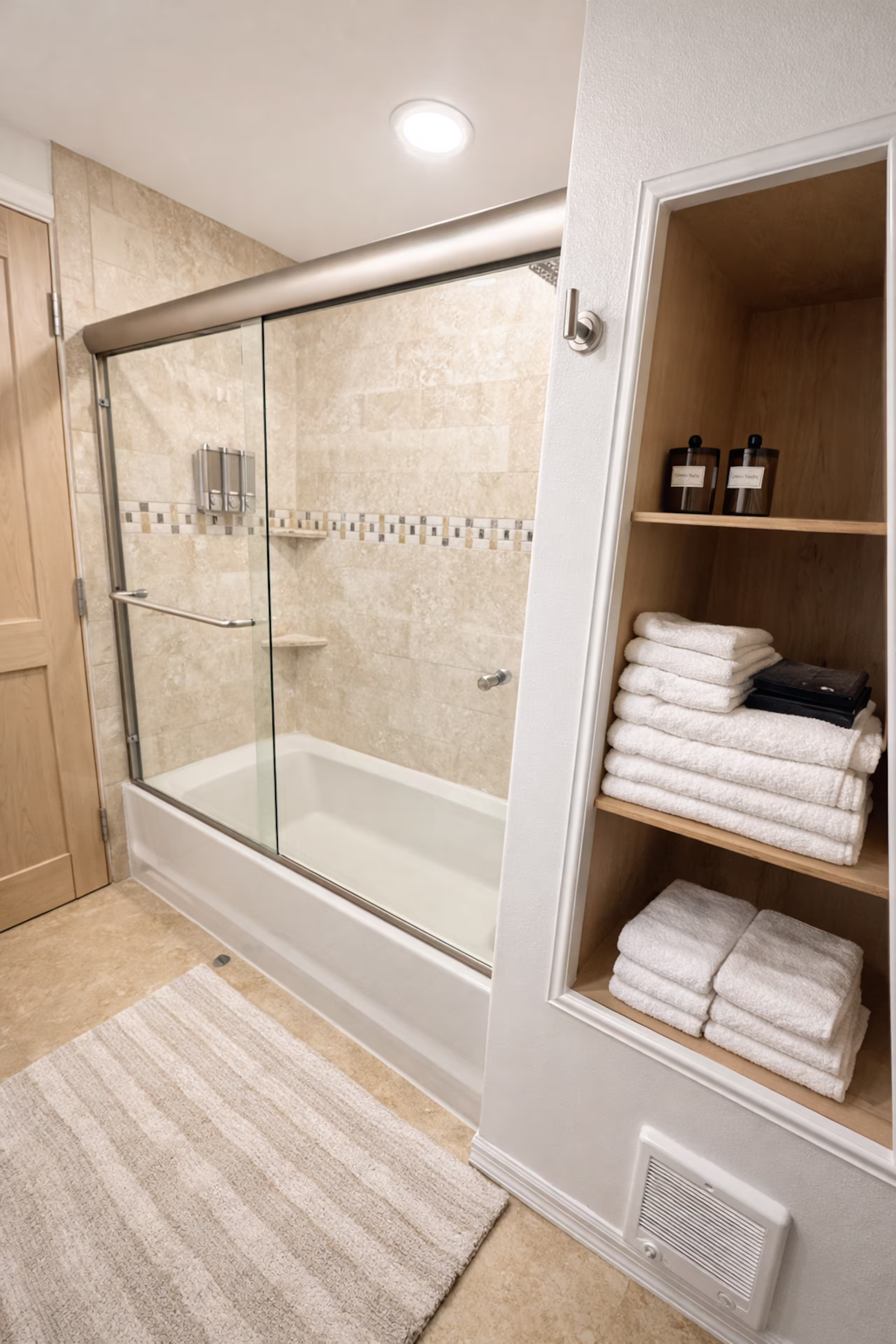 A bathroom shower with glass sliding doors, beige tiled walls, and a grab bar. Next to the shower is a built-in wooden shelf with folded white towels and brown bottles, and a beige and cream striped rug on the floor.