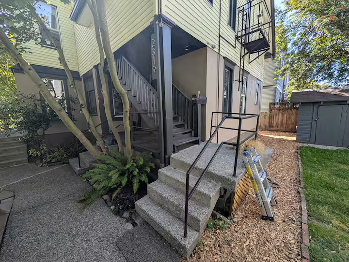 Exterior view of a multi-story house with a concrete staircase leading to the front door, black metal railings, a small garden area with plants, trees, and a lawn, and a ladder leaning against the house.