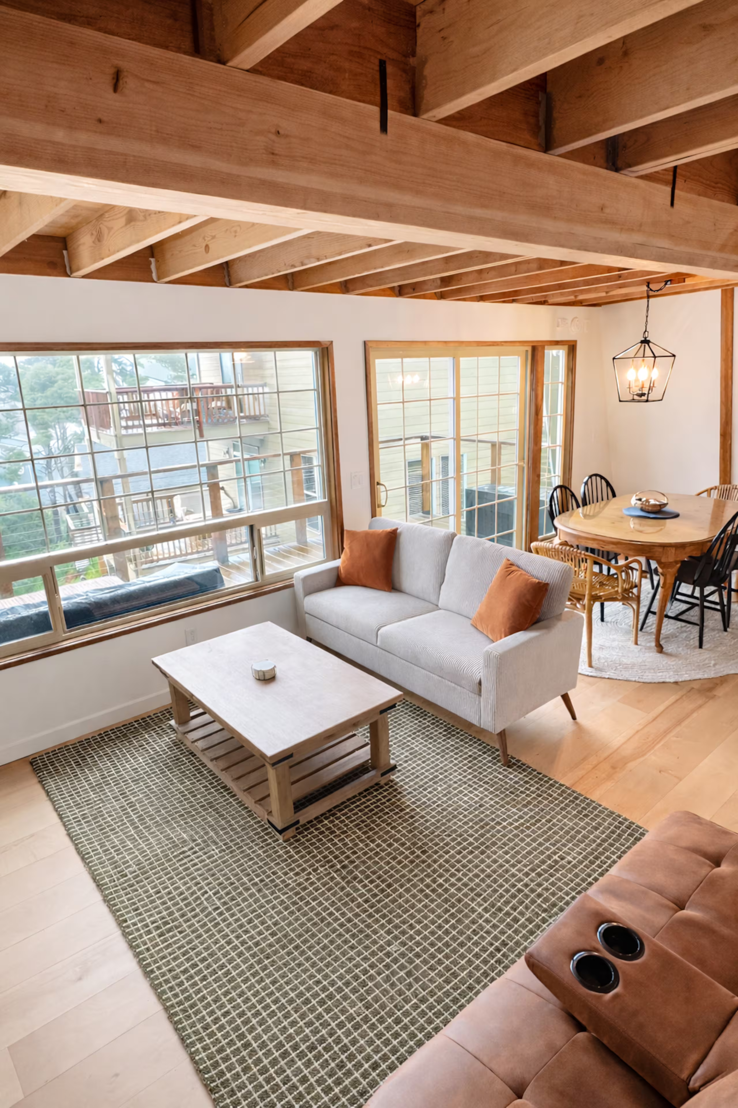 Living room with large windows, a white sofa with orange pillows, a wooden coffee table, a rug, dining table with chairs, and a chandelier. Exposed wooden ceiling beams.