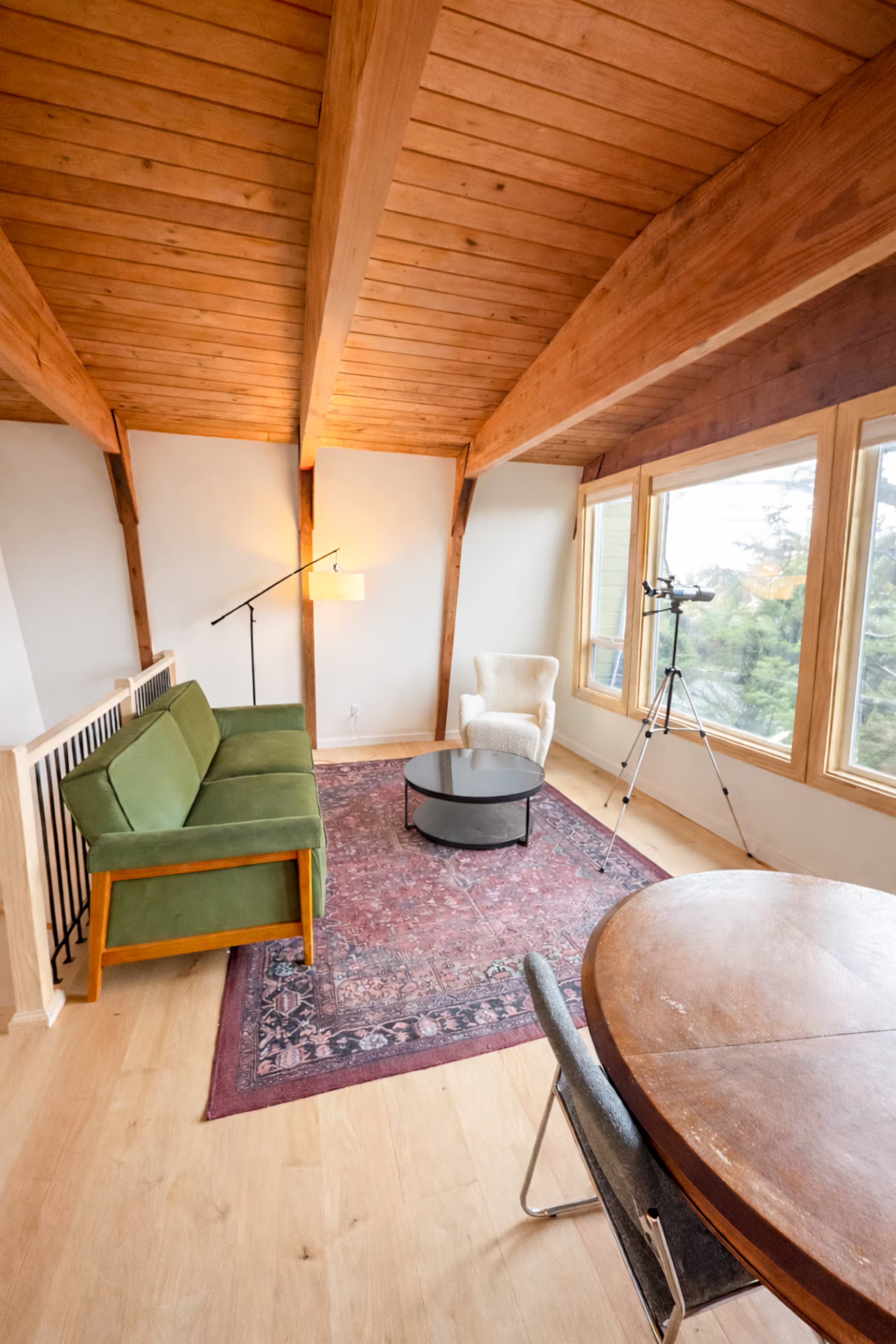 Living room with wooden vaulted ceiling, large windows, green armchair, white armchair, black coffee table, floor lamp, telescope near window, patterned area rug, and wooden dining table with chairs.