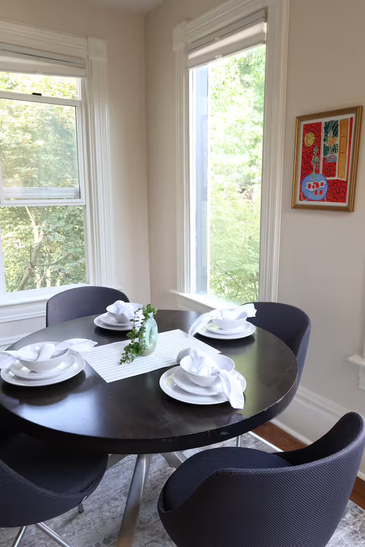 A round black dining table set for four, with white plates, napkins, and a small green vase with greenery, in a room with large windows and white walls, decorated with colorful modern art.