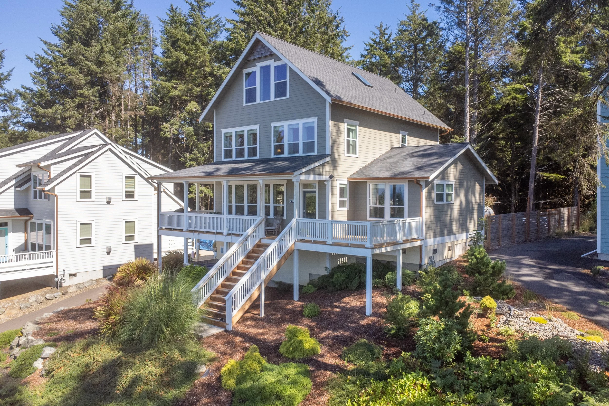 A modern multi-story house with gray siding, multiple large windows, and a screened porch with stairs leading down to landscaped yard. Tall evergreen trees in the background.