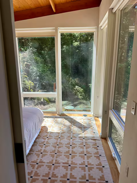 Sunlit bedroom with a sliding glass door and large window overlooking a lush outdoor scene, a bed with a white comforter on the left, and a patterned rug on the floor.