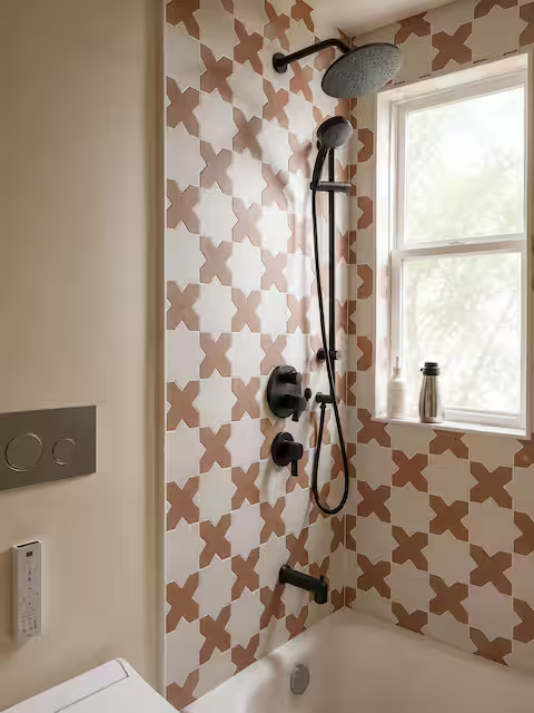 Bathroom shower area with brown and white geometric patterned tiles, a black showerhead and fixtures, a window with a bottle on the sill, and a partial view of a white bathtub.
