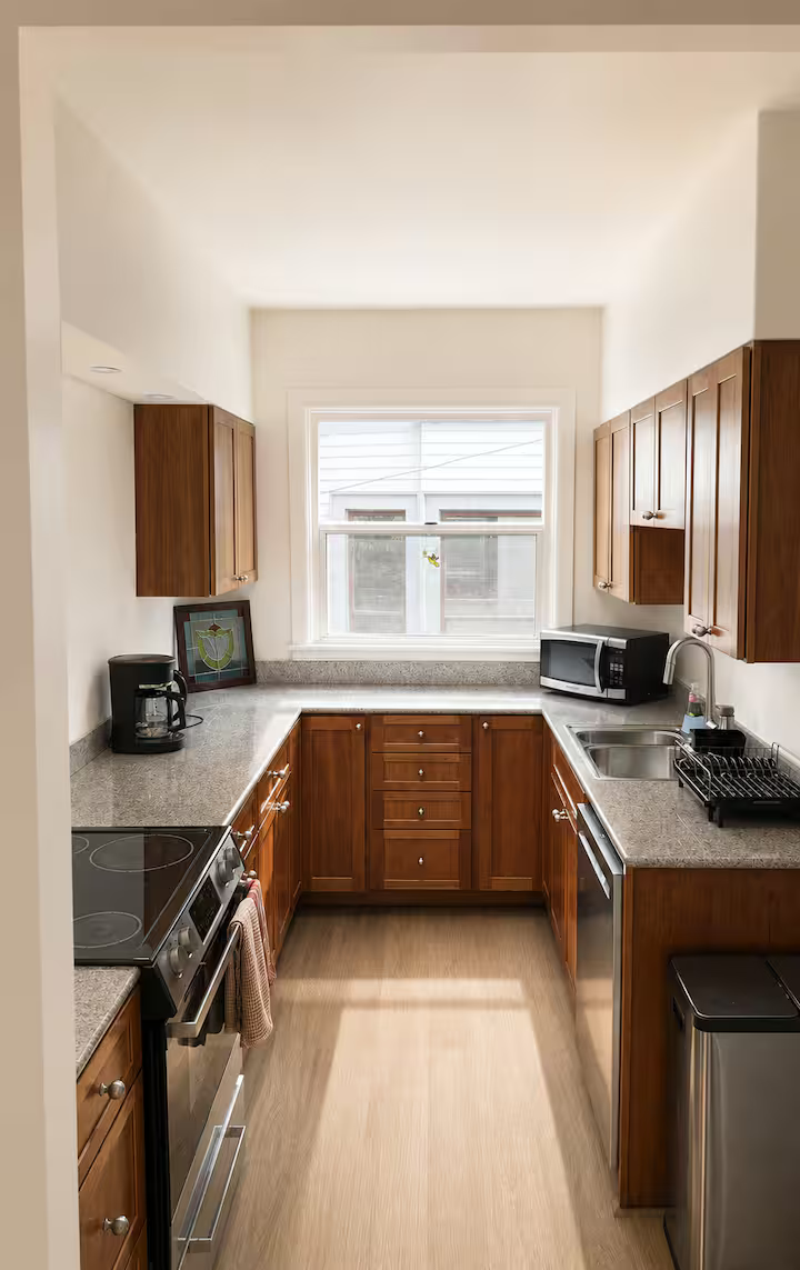 Small kitchen with wood cabinets, gray countertops, a window, microwave, coffee maker, stove, and sink.