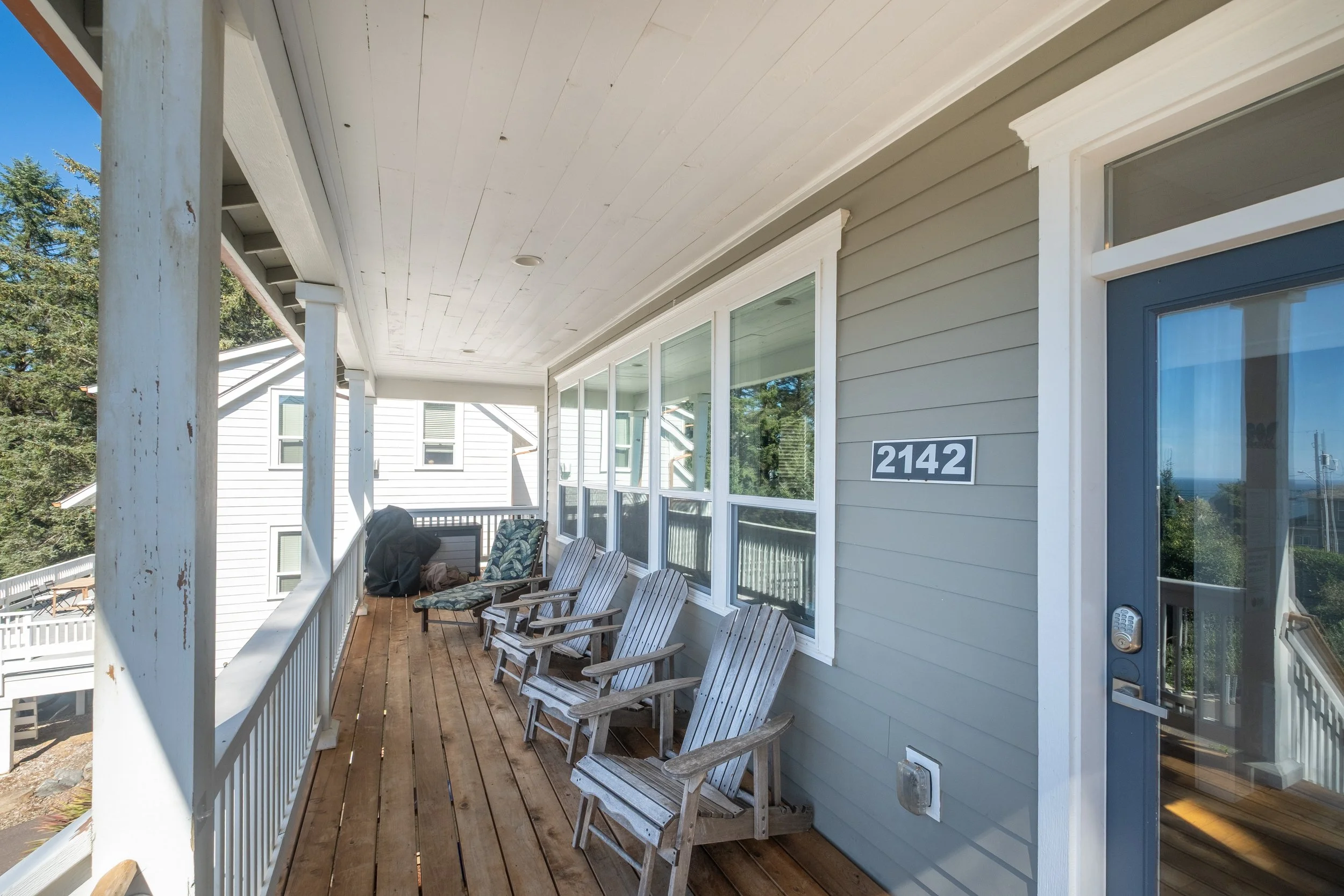 Front porch with wooden floor and railing, several wooden chairs, beige house siding, white trim, and glass door with the house number 2142, with trees and neighboring houses in the background under a sunny sky.