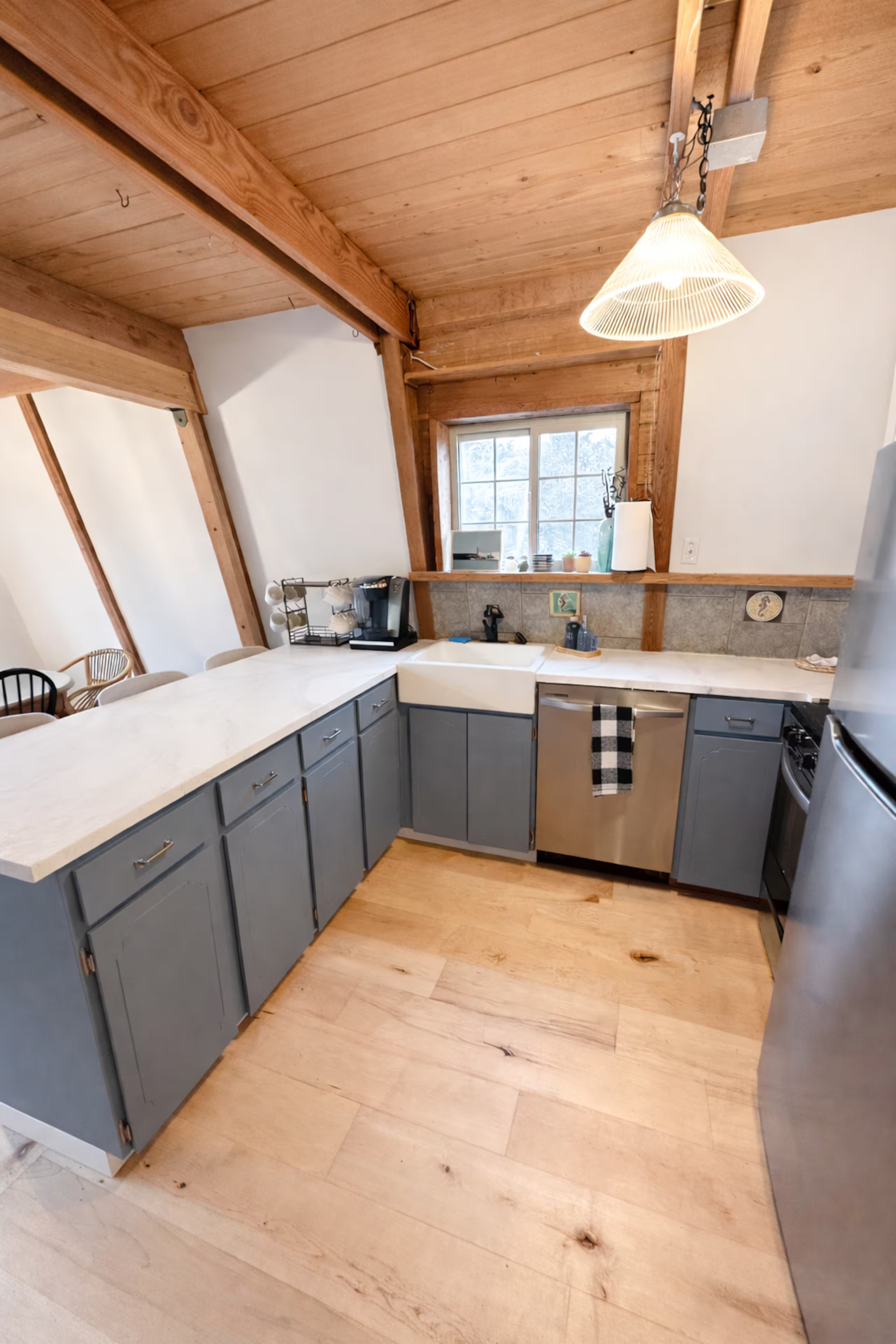 Kitchen with wooden ceiling and cabinets, gray lower cabinets, white countertops, window above the sink, various small appliances and decor on the counter, and a light fixture hanging from the ceiling.