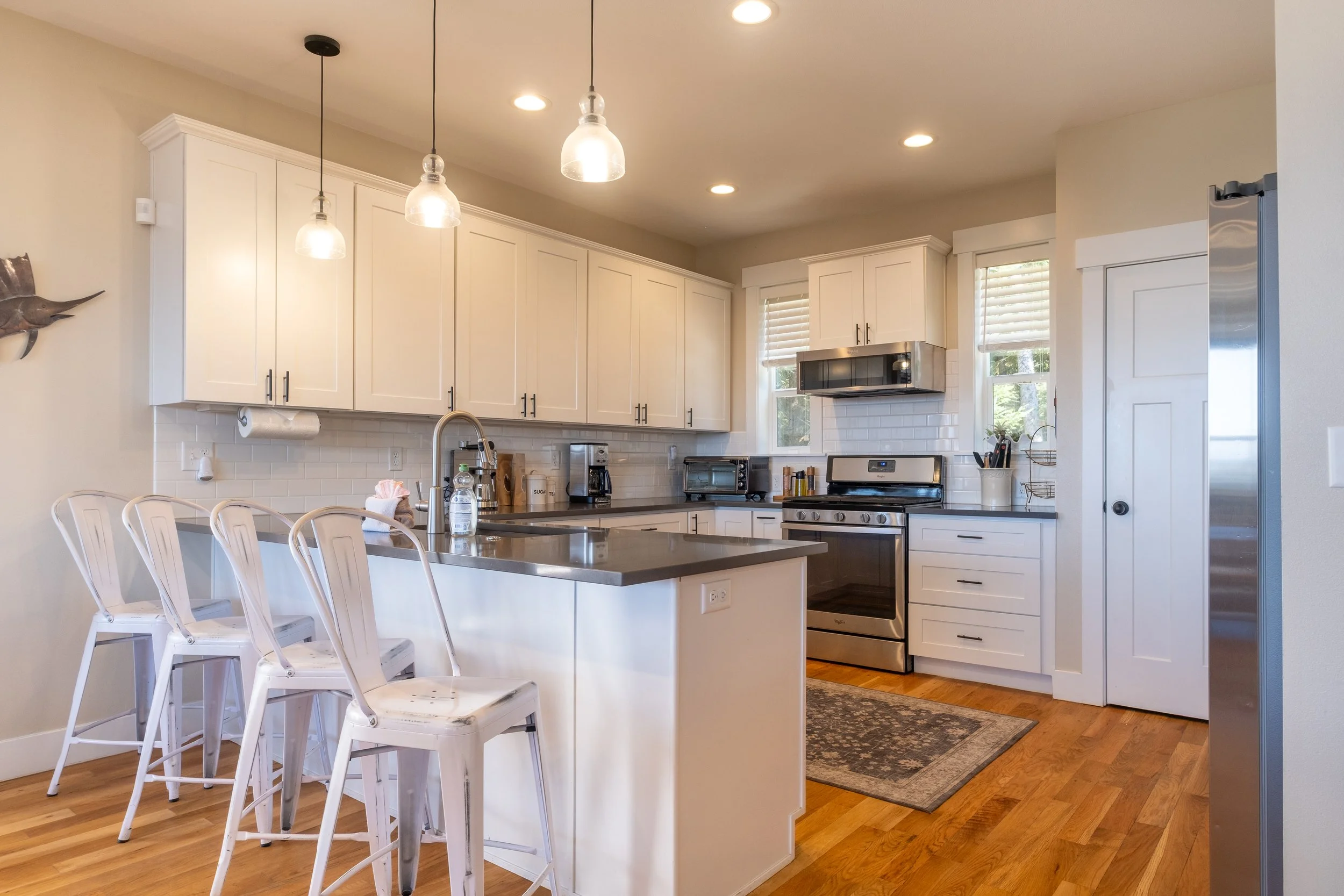 A modern kitchen with white cabinets, black hardware, stainless steel appliances, a kitchen island with white chairs, pendant lights, and windows with white blinds.