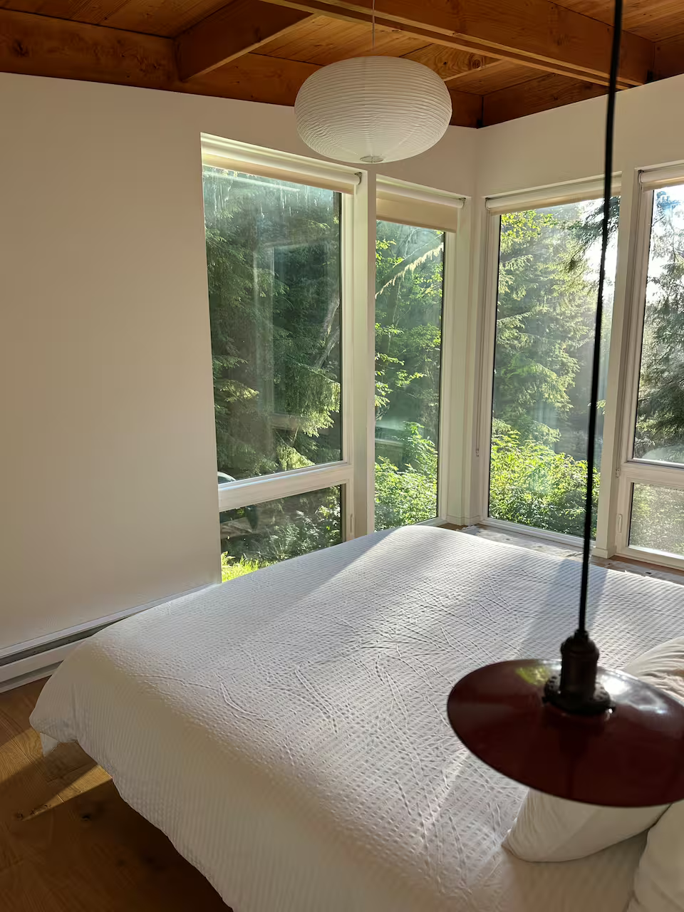 A bedroom with large windows showing green trees outside. The bed has a white quilt and pillow, with a pendant light hanging from the ceiling.