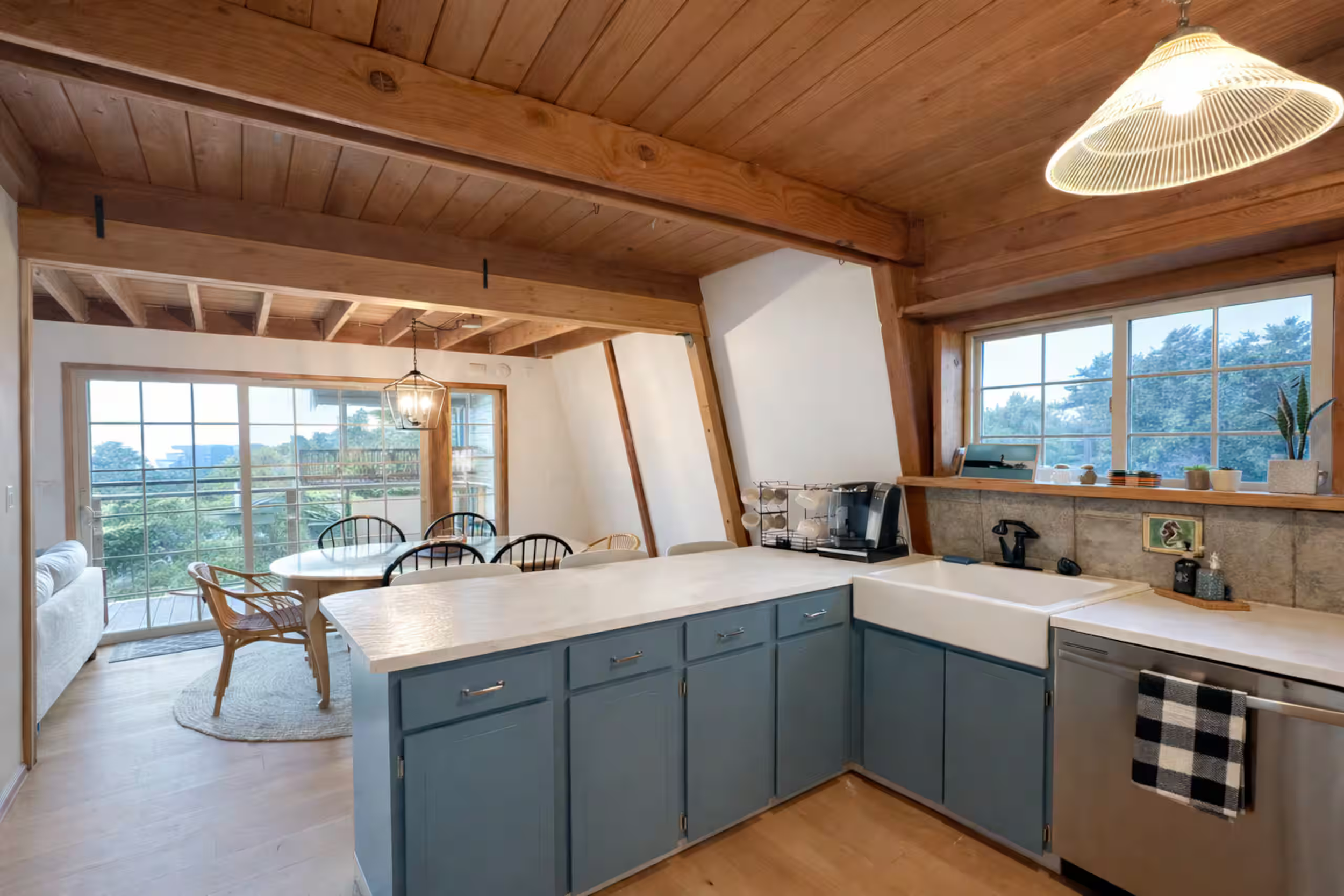 Open-concept living space with a kitchen and dining area featuring a wooden ceiling, large windows, and a view of trees outside. Kitchen has blue cabinets, white countertops, a white farmhouse sink, and various small appliances. Dining table with cha