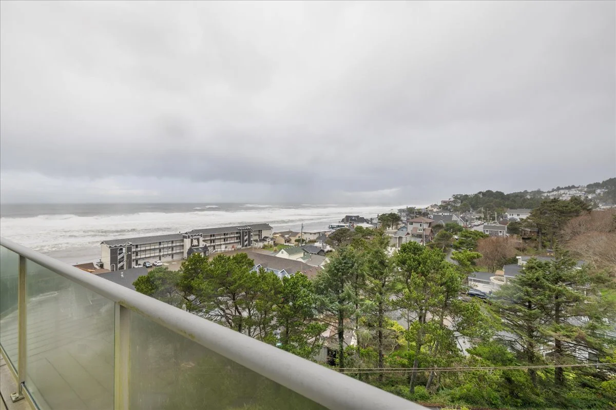 View from a balcony overlooking a coastal neighborhood with trees and houses, ocean with waves crashing on the shore, overcast sky.