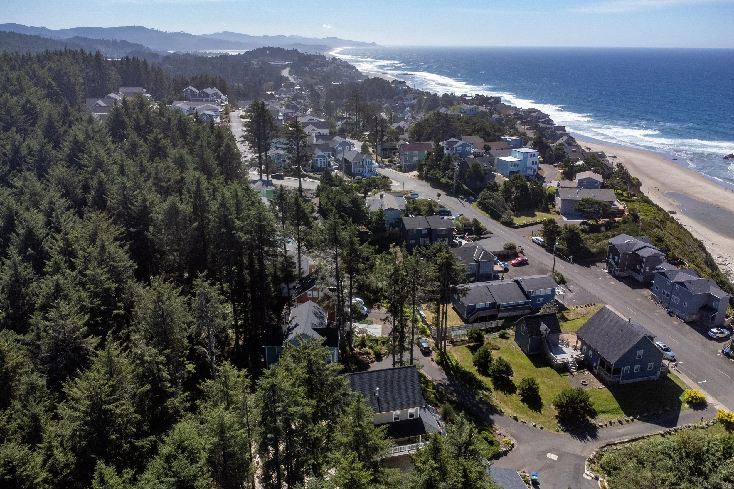 Aerial view of a coastal neighborhood with houses along the ocean, surrounded by forested hills and mountains.