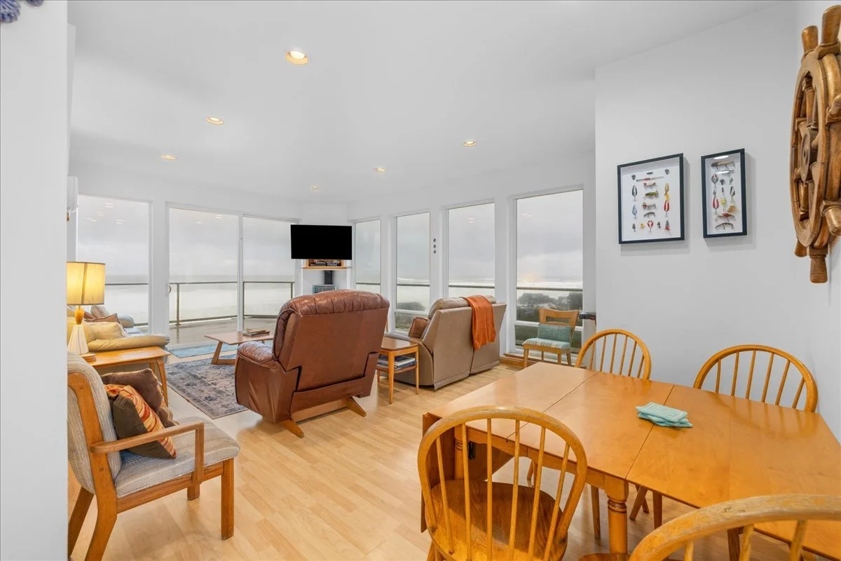 Living room with large windows, wooden dining table, leather and fabric chairs, a television, and corner with ocean view.