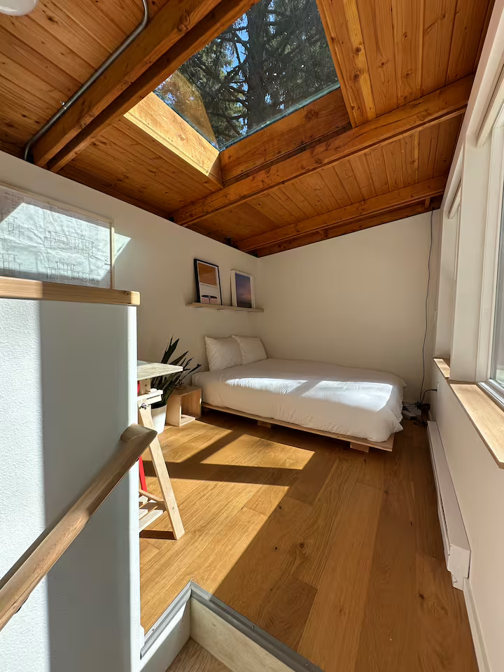 Sunlit bedroom with a sloped wooden ceiling and a skylight, white bed with pillows, wooden floor, window with a view of trees, and small decorative pictures on a shelf.