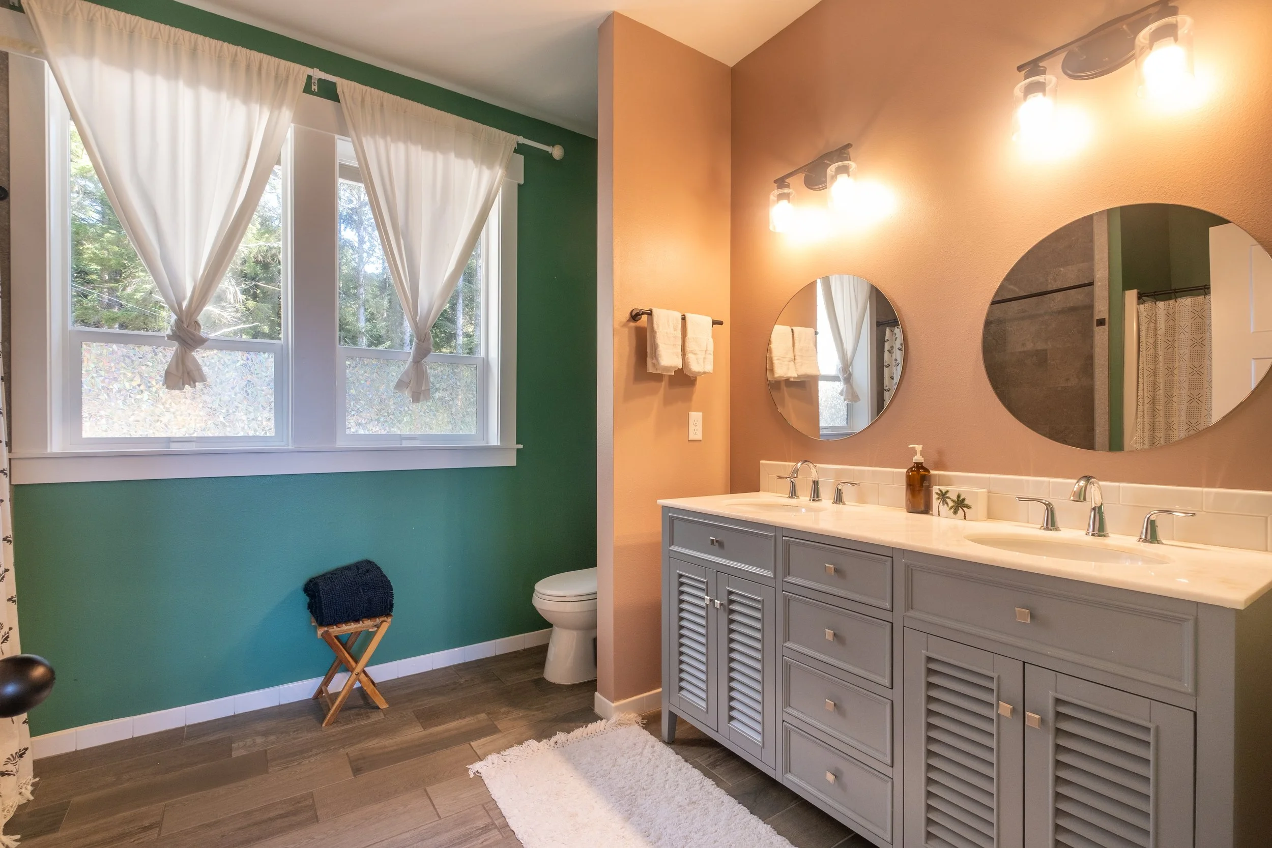 Bathroom with a double sink vanity, round mirrors, a beige wall, and a window with white curtains.