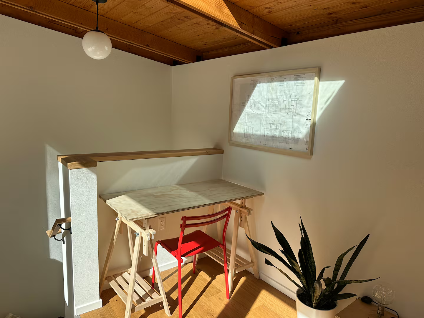 A small home-office corner with a wooden drafting table, a red chair, a potted snake plant, a window with sunlight, and a framed architectural blueprint on the wall.