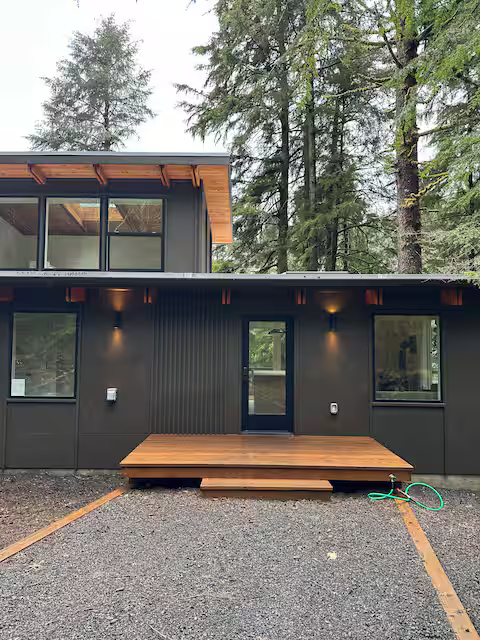 Modern black house with wooden porch, surrounded by tall pine trees in a wooded area.