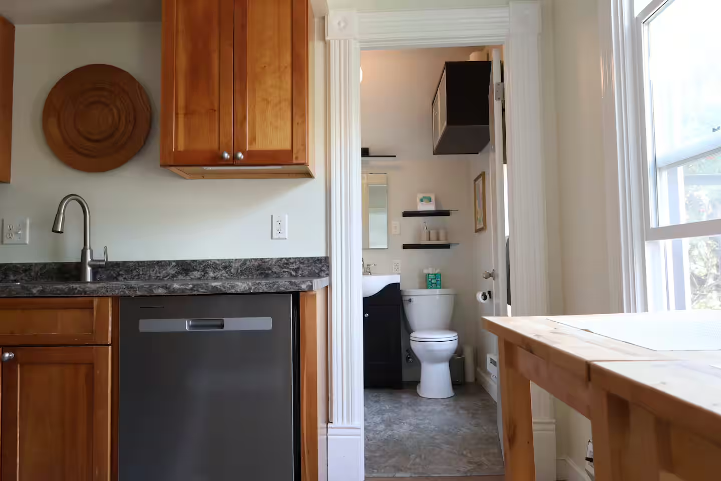 Interior view of a kitchen leading to a small bathroom with a toilet, shelves, and window, featuring wooden cabinets, black countertop, and a dishwasher.