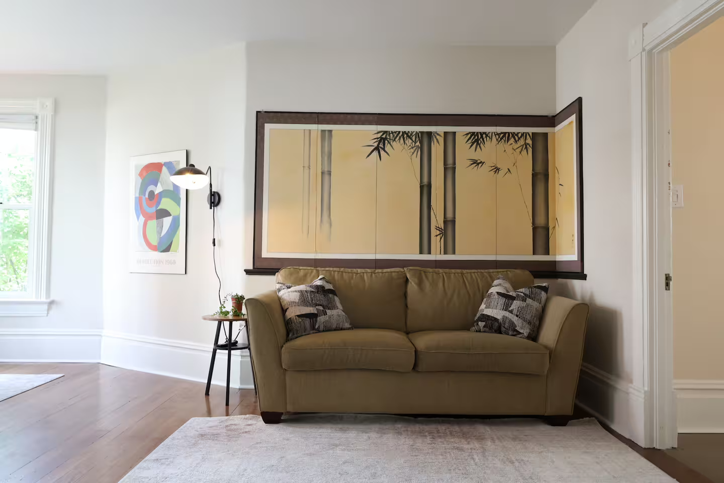 Living room interior with beige sofa, colorful pillows, large bamboo wall art, side table with plant, wall-mounted lamp, and abstract art on white walls, wood flooring, and window with trees outside.