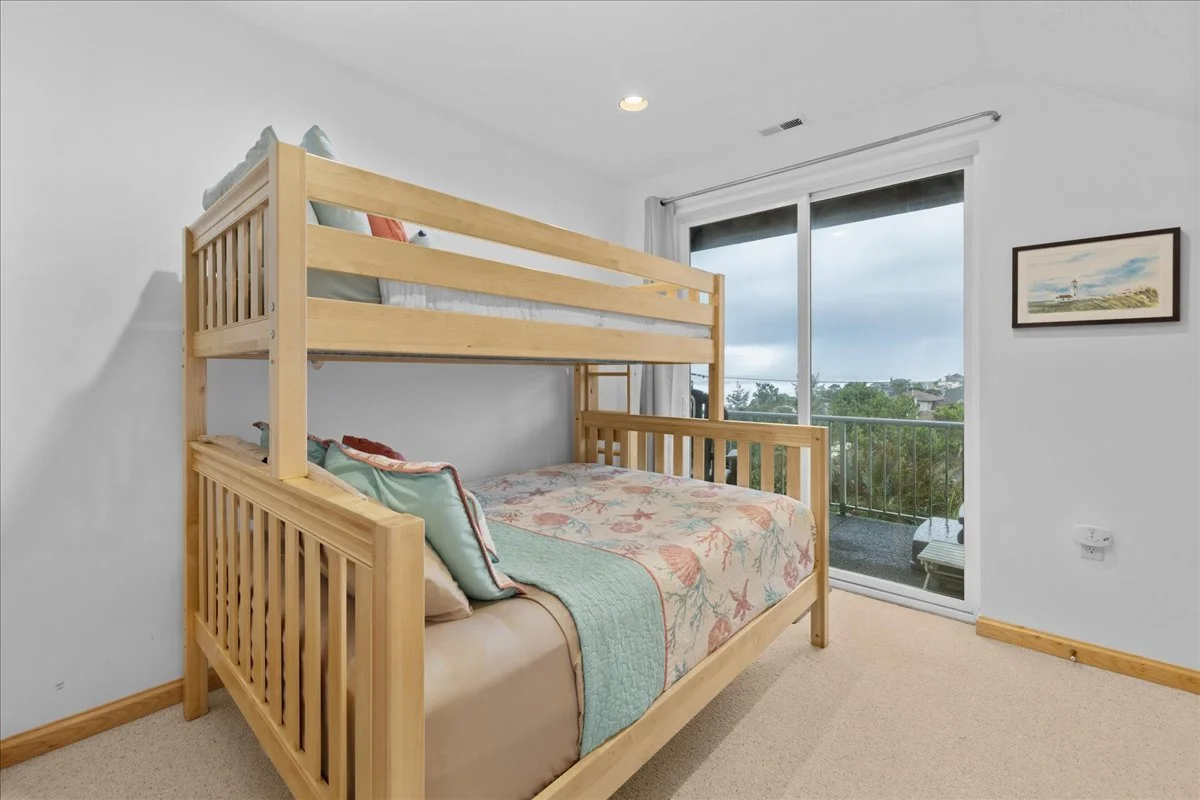 A bedroom with a wooden bunk bed, featuring a lower bed and an upper bed. The lower bed has a floral quilt and pillows, while the upper bed has pillows. There is a sliding glass door leading to a balcony with outdoor furniture, and a framed picture o