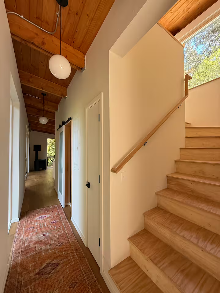 Interior view of a hallway in a house, featuring wooden stairs, a door with a black handle, a carpet runner, and a vaulted ceiling with wood paneling. Sunlight coming through a window illuminates the space.